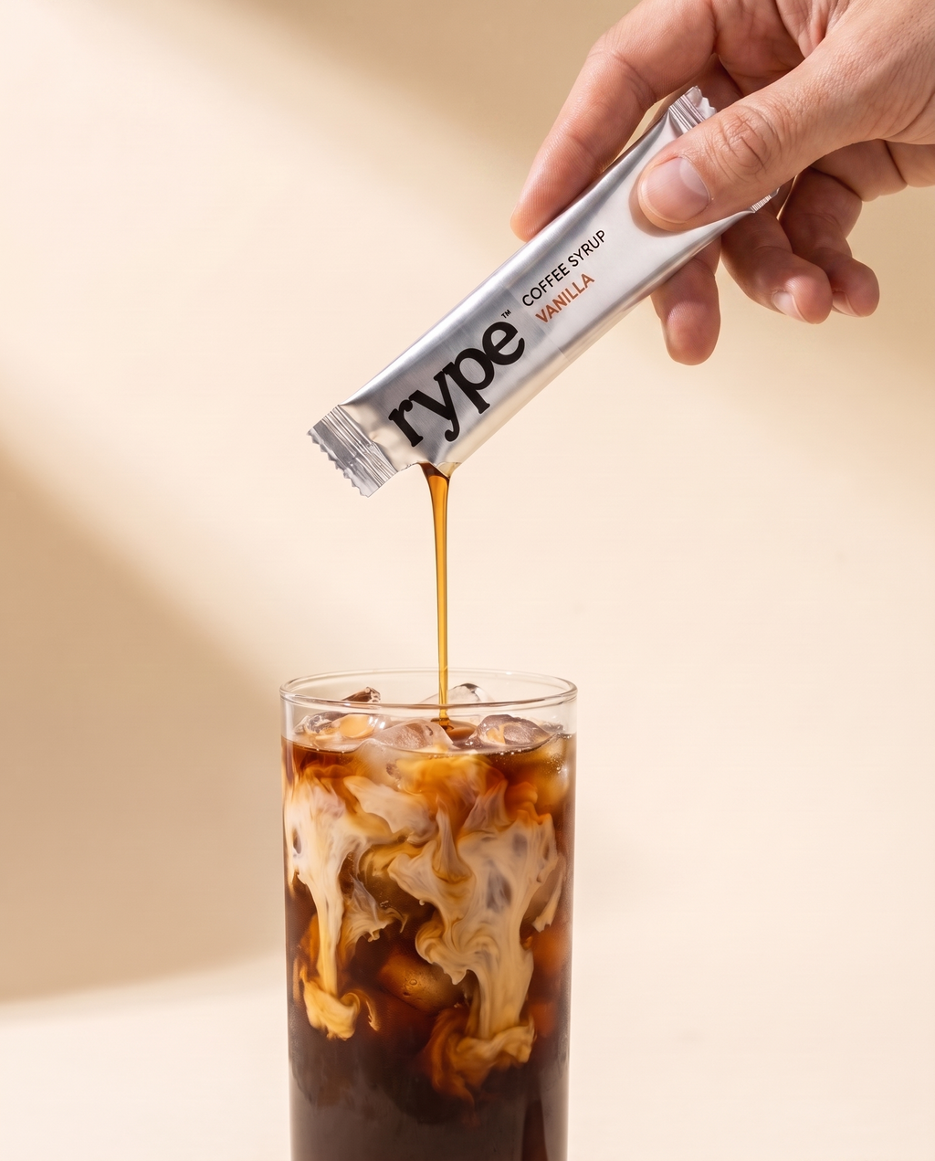 A hand tipping a chrome Rype sachet, pouring vanilla coffee syrup into a tall glass of iced coffee